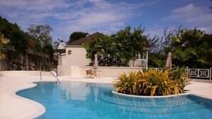 A swimming pool with a curved edge, surrounded by tropical plants and a white deck. - Apartment with Pool set In 70 Acres Of Beautifully Lush Tropical Gardens (Rockley)