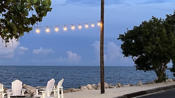 A beachside setting with white Adirondack chairs, a string of lights, and a view of the ocean.