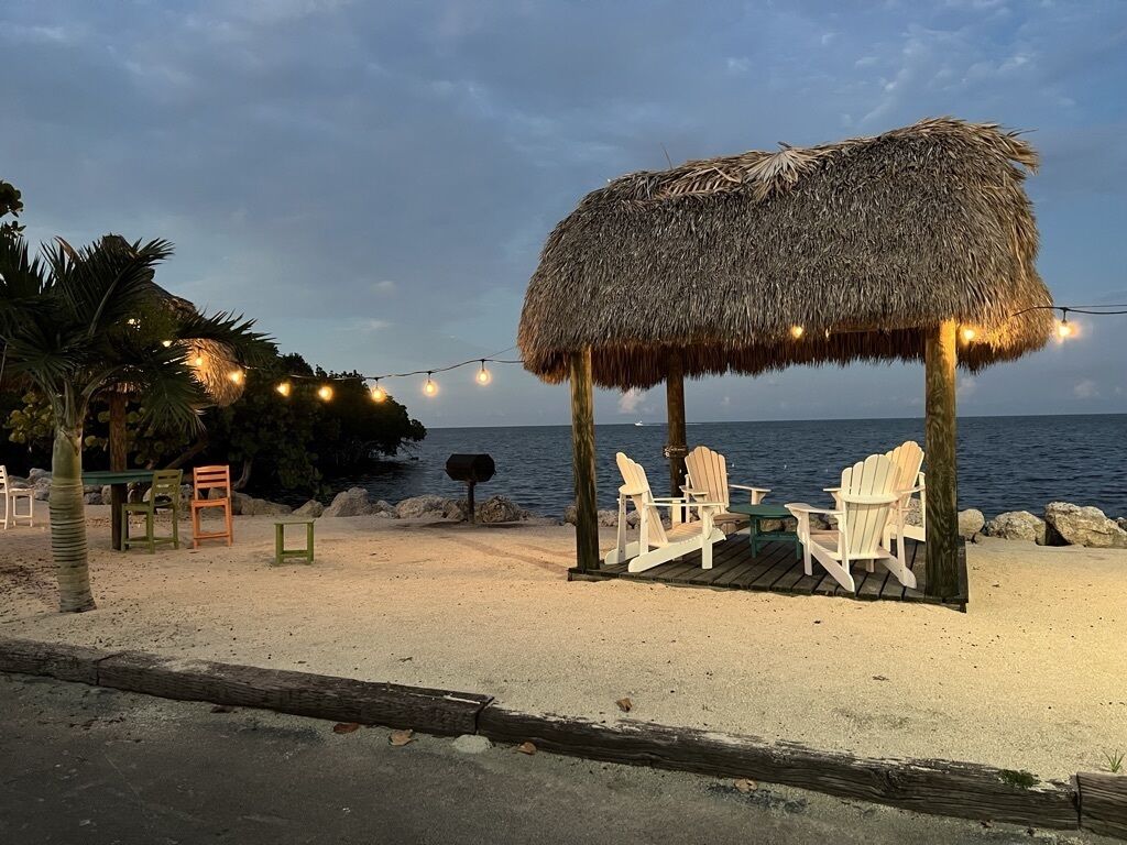 A beachside seating area with a thatched roof, white Adirondack chairs, and string lights.