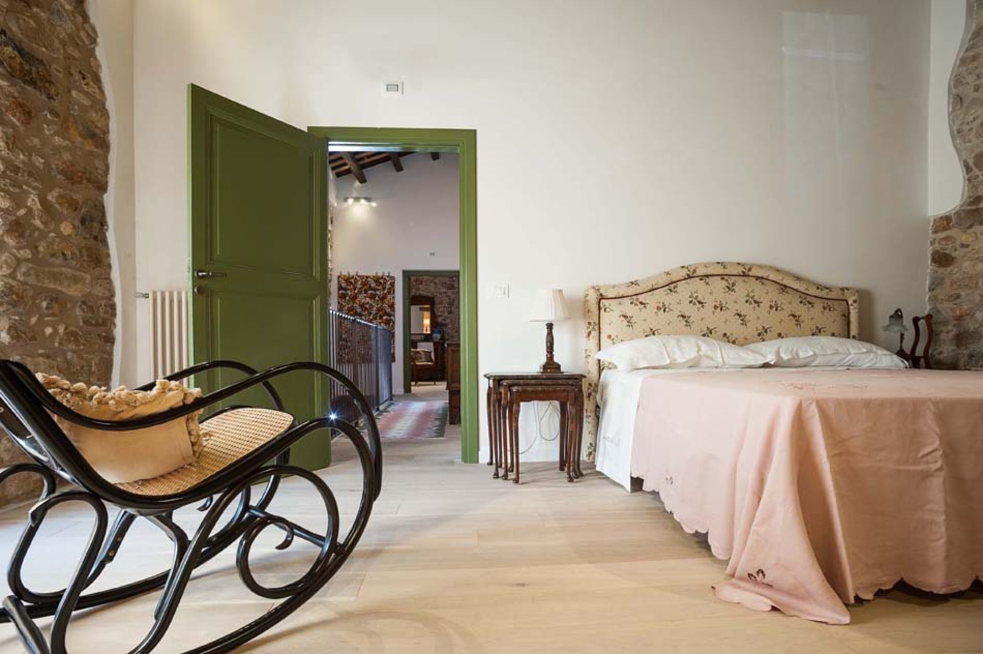 Um quarto com piso de madeira, porta verde, uma cadeira de balanço, cama com cabeceira estampada e uma mesinha de cabeceira.