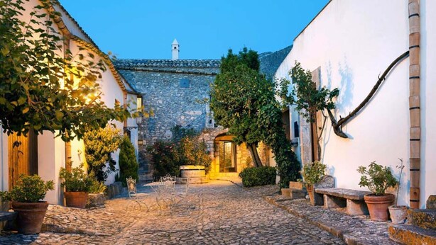 A cobblestone street in a historic town with white buildings, stone walls, and potted plants. - Holiday Home in Sicily for 6 People with Pool (Buseto Palizzolo)