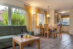 Ett mysigt vardagsrum med en grön soffa, ett matbord i trä och stolar. Det finns ett fönster med gardiner, en lampa och en utsikt över grönskan utanför.