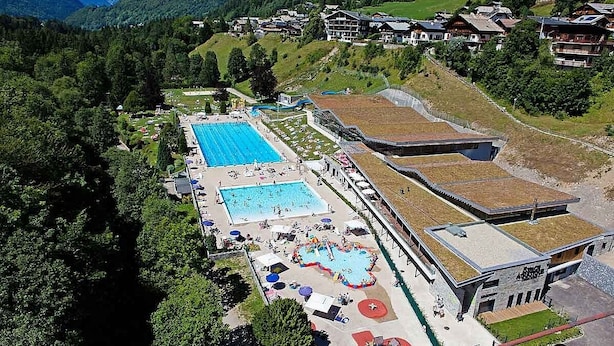 An aerial view of a resort with multiple swimming pools, a large building with a green roof, and a mountainous backdrop. - Unique, charming chalet with ensuite rooms, hot tub and games room (Saint-Jean-d'Aulps)