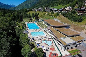 Vista aérea de un complejo turístico con varias piscinas, un edificio grande con techo verde y montañas al fondo.