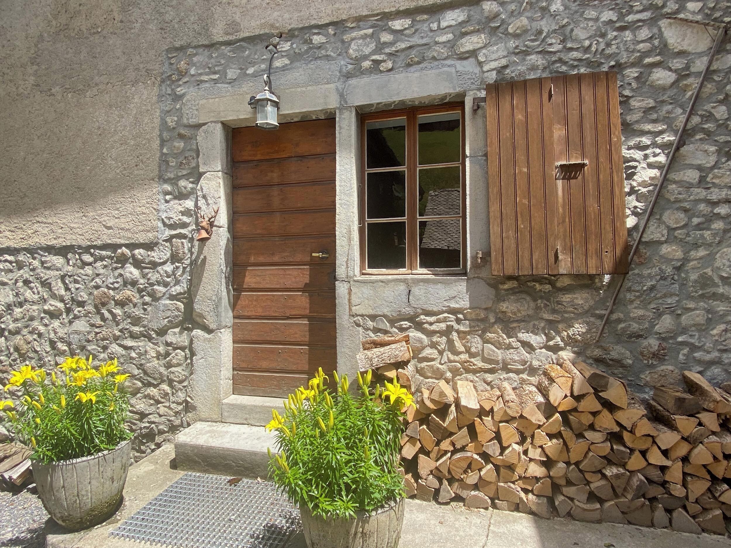 Un bâtiment rustique en pierre, doté d’une porte en bois, d’une fenêtre avec des volets en bois et d’une lanterne au-dessus de la porte. On y trouve également des plantes en pot et une pile de bois de chauffage à l’extérieur.
