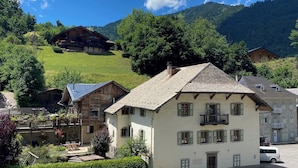 A traditional chalet-style building with a sloped roof and wooden exterior, surrounded by lush greenery and a mountainous backdrop. - Unique, charming chalet with ensuite rooms, hot tub and games room (Saint-Jean-d'Aulps)