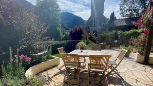 A stone patio with a wooden table and chairs, surrounded by lush greenery and flowering plants. - Unique, charming chalet with ensuite rooms, hot tub and games room (Saint-Jean-d'Aulps)