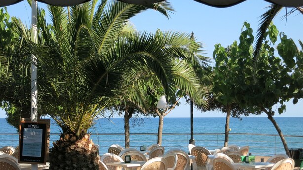 A beachside restaurant with outdoor seating, a menu board, and a view of the sea.