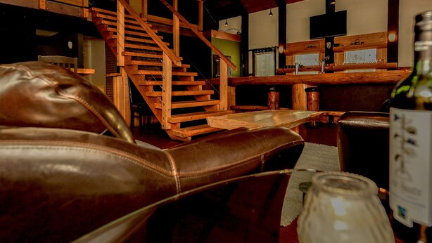 A cozy lounge area with leather seating, a wooden staircase, and a bar area with bottles and glasses. - One Of A Kind Vacation Rental Located By The Base Of Boulder Mountain! (Columbia-Shuswap B)