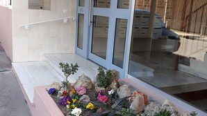 A building entrance with a glass door, a small flower bed with various plants, and a metal signboard on the wall. - GOLFE JUAN, 150 m from the beach, F2, bright, 43m2, balcony 14m2, sea view + garage (Vallauris)