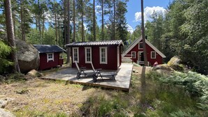Une cabane dotée d’une terrasse en bois et de deux chaises, entourée de grands arbres et d’un gros rocher.