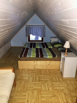 Un pequeño dormitorio en el ático con techo inclinado, una cama con colcha a rayas, una mesita de noche con lámpara y una ventana con cortina a cuadros.
