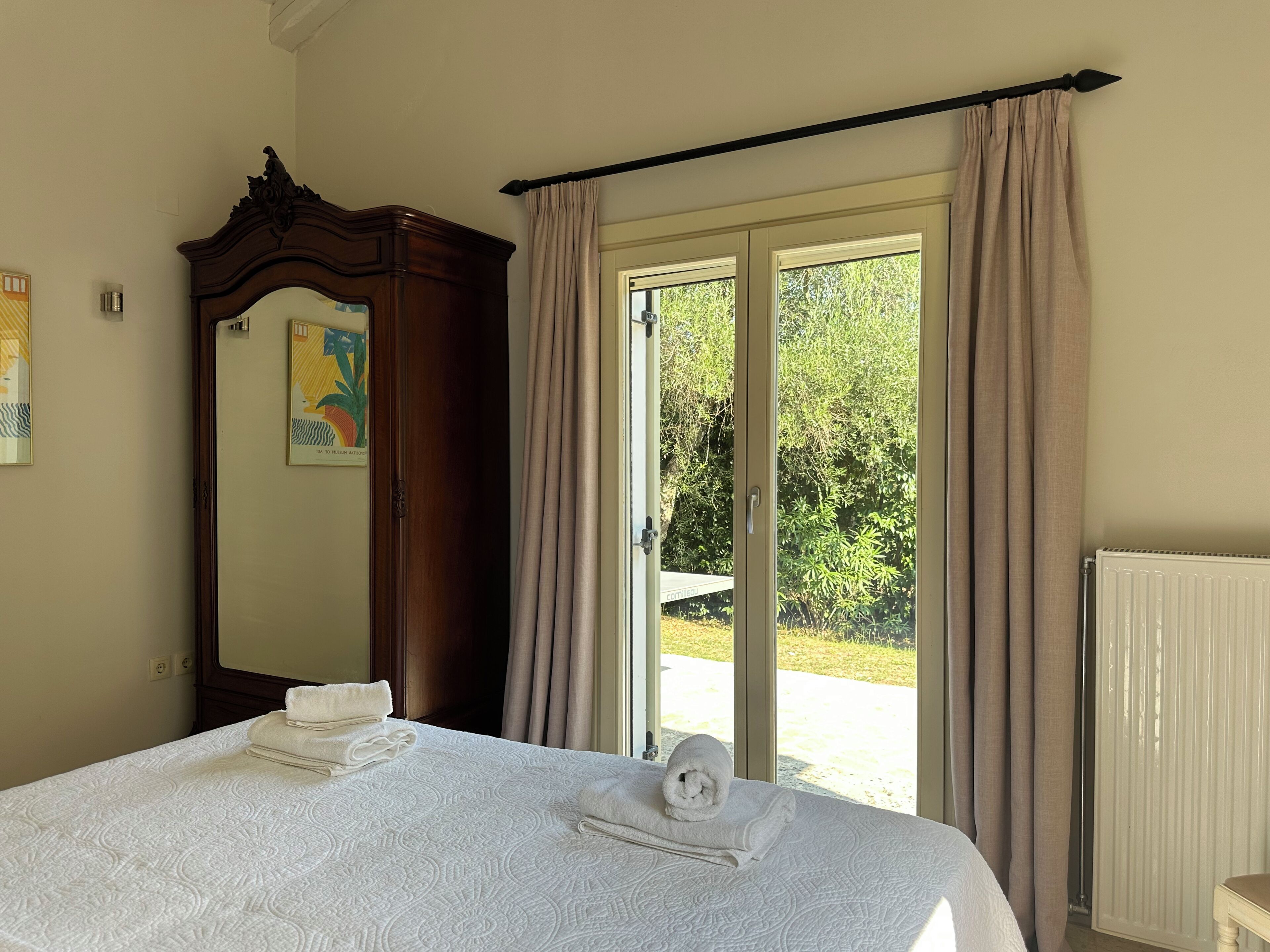 Een slaapkamer met een bed, een houten garderobe, een spiegel en uitzicht op groen via een glazen schuifdeur.