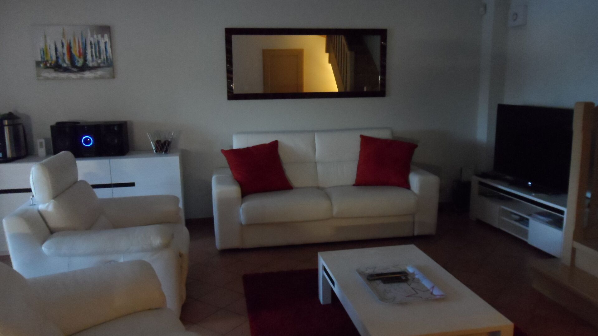 Una sala de estar con un sofá blanco, cojines rojos, una mesa de centro blanca, un televisor de pantalla plana y un cuadro en la pared.