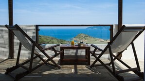 A balcony with two white canvas chairs and a small wooden table with a glass of juice, overlooking a coastal landscape.