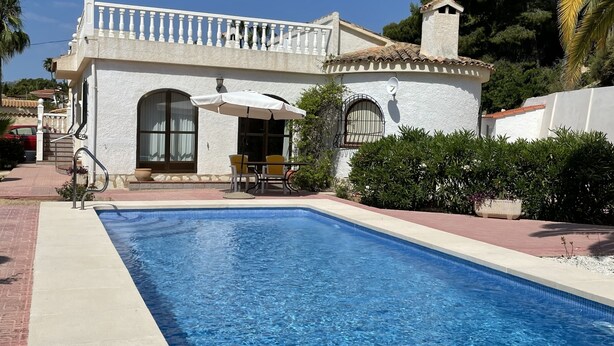 Zona de piscina con una pileta azul cristalina, un edificio blanco con balcón y una palmera al fondo.