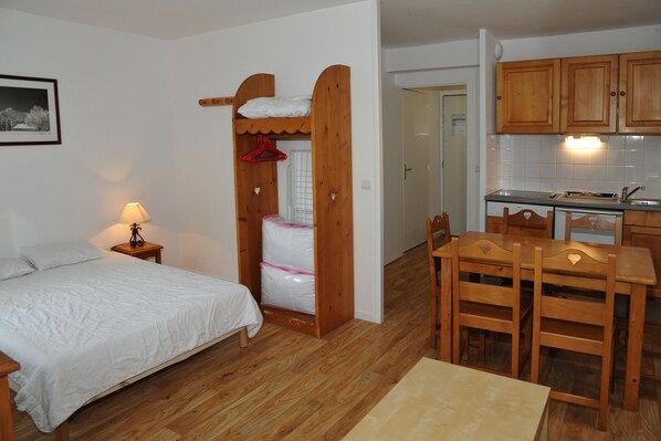 Et lille, funktionelt værelse med seng, et lille bord, træstole, et tekøkken og et træskab.