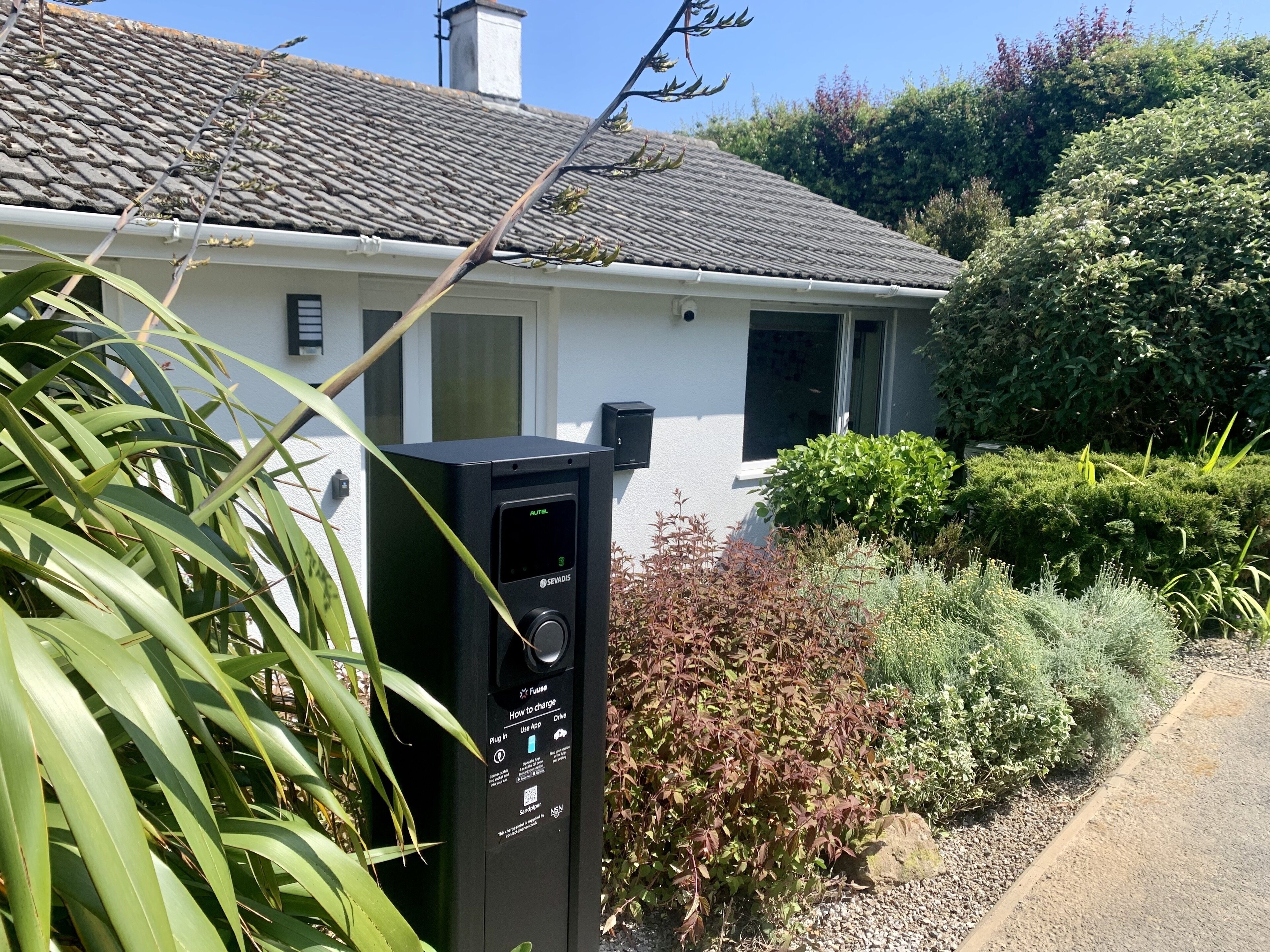 Une borne de recharge pour véhicules électriques, de couleur noire, avec des instructions, située devant une maison dont le toit est recouvert de tuiles et qui est entourée de verdure.