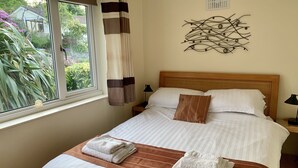 Een netjes opgemaakt bed met witte lakens en kussens met bruine strepen, een raam met gordijnen, en een abstract kunstwerk aan de muur.