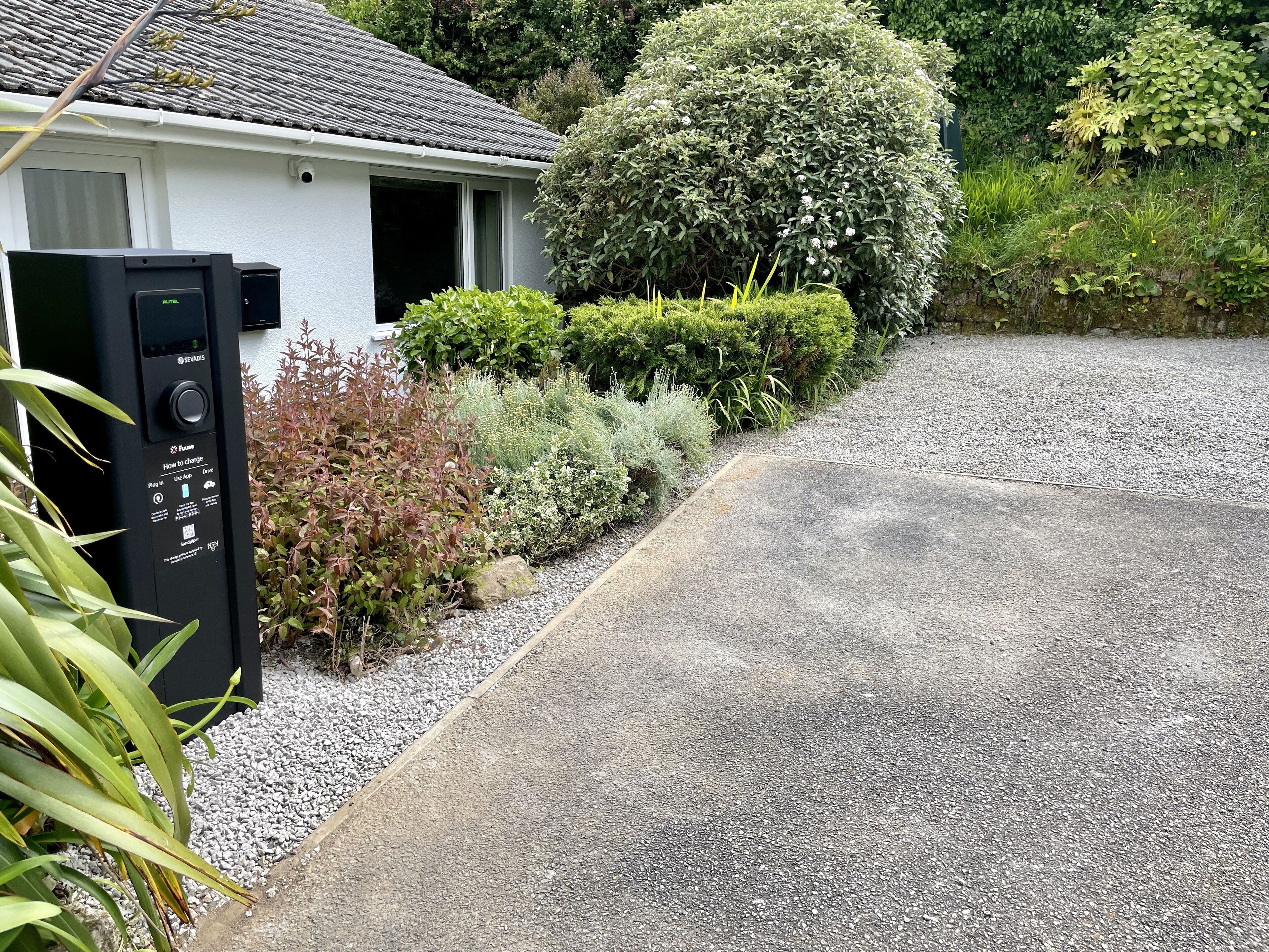 A residential driveway with a charging station, a white house, and a well-maintained garden.