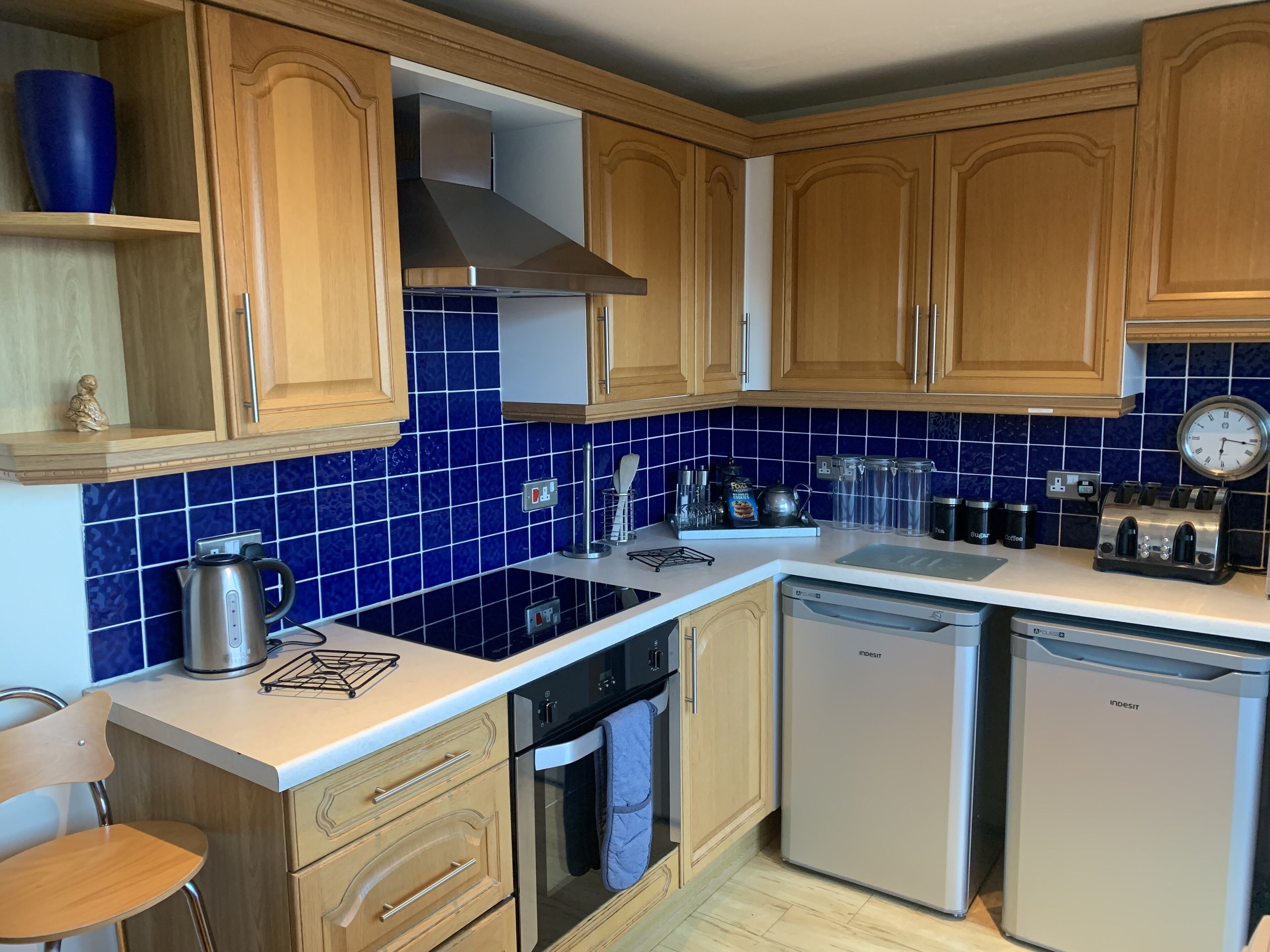 Una cocina con armarios de madera, salpicadero de azulejos azules, electrodomésticos de acero inoxidable y un reloj en la pared.