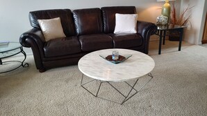Un canapé en cuir marron foncé agrémenté de deux coussins blancs, une table basse ronde avec un plateau en verre et une petite table d’appoint sur laquelle sont posés une lampe et quelques objets décoratifs.