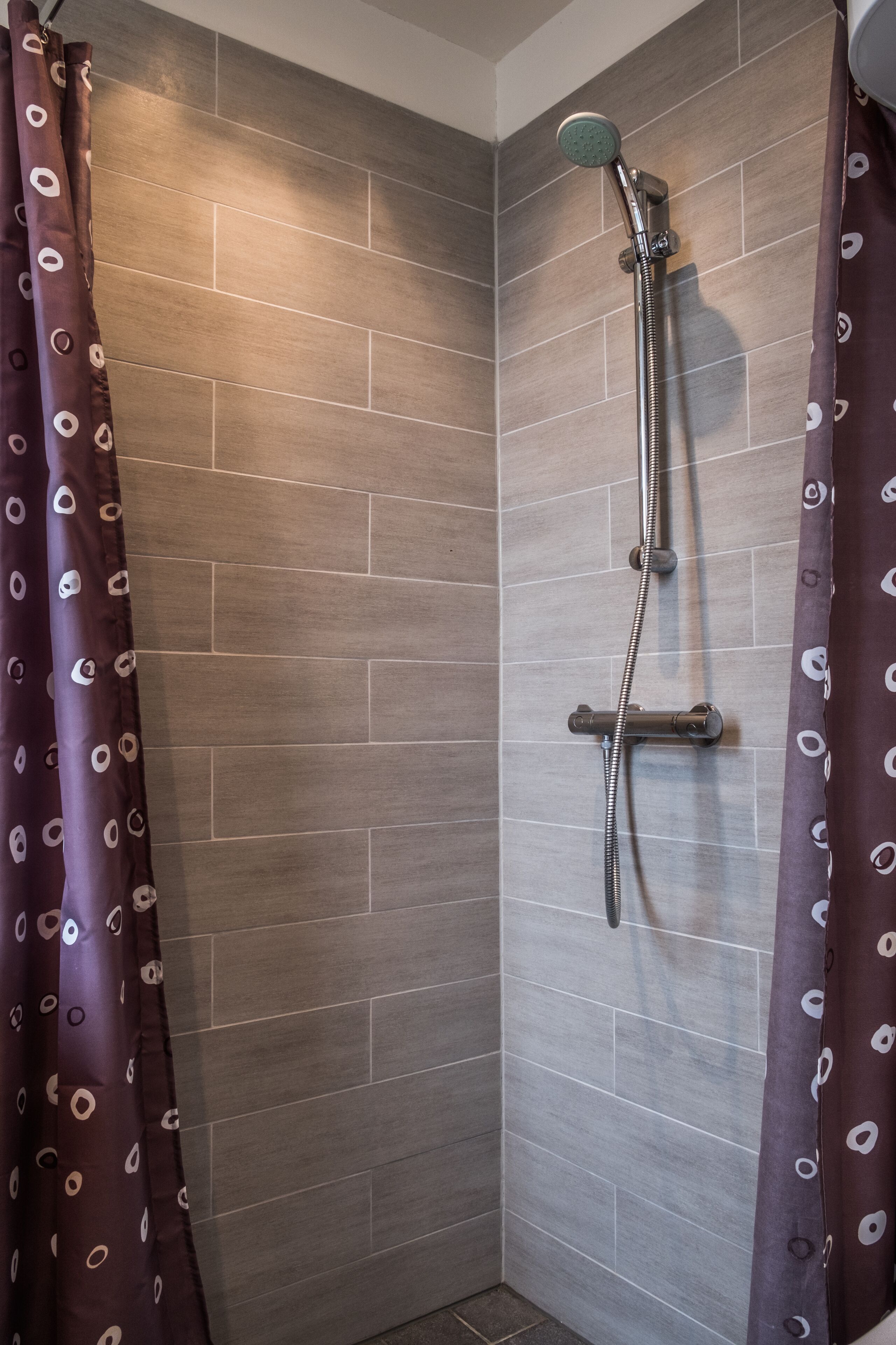 A shower with a handheld showerhead and a wall-mounted faucet, enclosed by a curtain with a pattern.