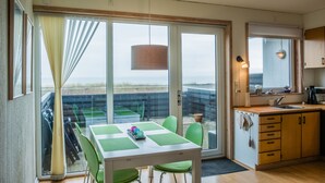 Ett rum med ett matbord, stolar, en köksdel och ett stort fönster med utsikt över en strand.
