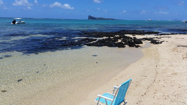 Una playa con aguas transparentes y poco profundas, una silla y una isla rocosa al fondo.