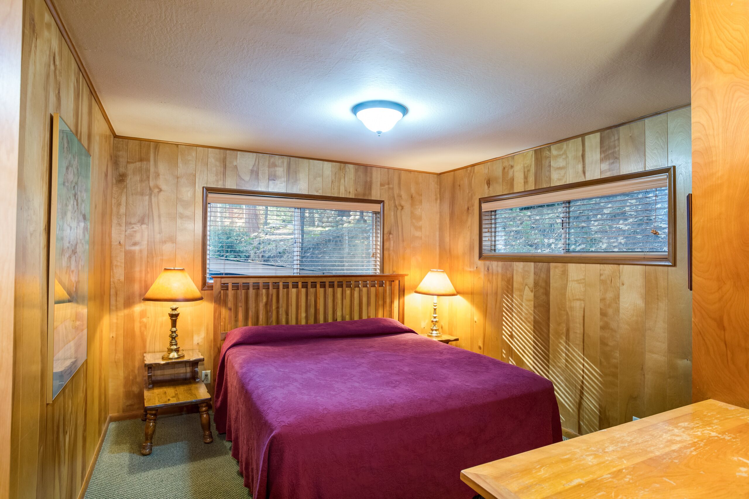 Una habitación con paneles de madera, una cama con colcha morada, dos lámparas de noche, una ventana con persianas y un cuadro en la pared.