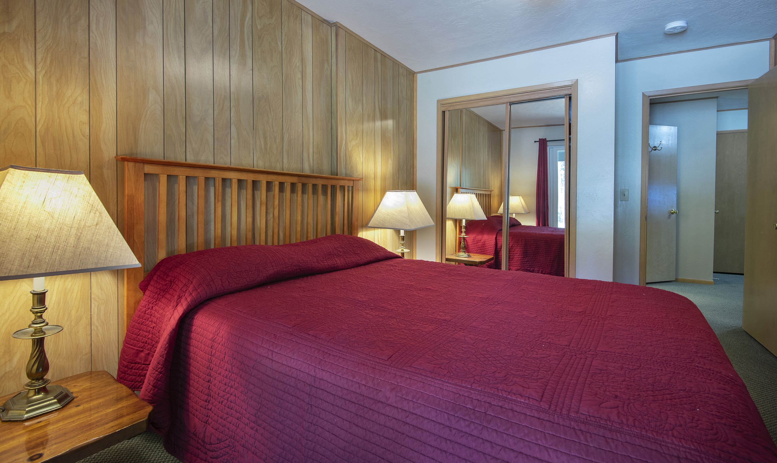 Ett hotellrum med en sänggavel i trä, en stor säng med ett rött täcke, två sänglampor och en spegel som reflekterar rummet.