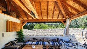 Un espace extérieur abrité, avec des bancs en bois et des coussins, une table en verre et un mur de pierre en arrière-plan.
