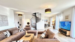 Ein modernes Wohnzimmer mit Wendeltreppe, einem großen Flachbildfernseher und einem Essbereich im Hintergrund.