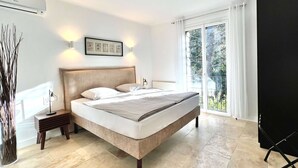 Ein Schlafzimmer mit einem Bett, einem Nachttisch, einem gerahmten Bild und Blick ins Freie durch eine Glastür.
