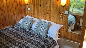 A wooden-paneled room with a bed, two wall-mounted lamps, and a mirror reflecting a window view. - KINGLAS CABIN is in a lovely, peaceful location deep in the forest, with hot tub (Dalavich)