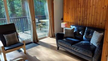 Ein gemütliches Blockhaus-Interieur mit einem Ledersofa, einem Holzstuhl und Blick auf eine Holzterrasse und Bäume draußen.