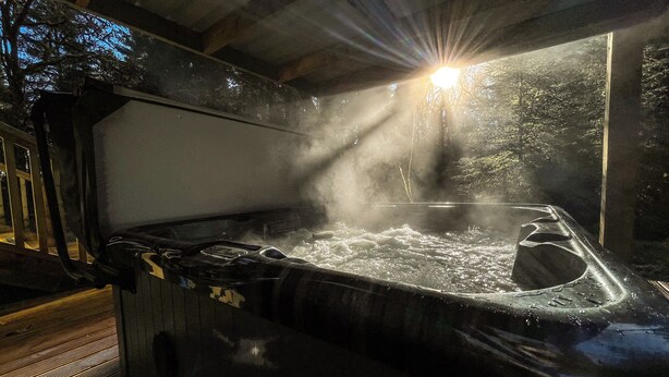 A hot tub with steam rising from the water, surrounded by a wooden deck and a metal railing. - KINGLAS CABIN is in a lovely, peaceful location deep in the forest, with hot tub (Dalavich)