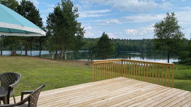 Una terraza de madera con vistas a un lago, rodeada de árboles, y una zona cubierta con sillas.