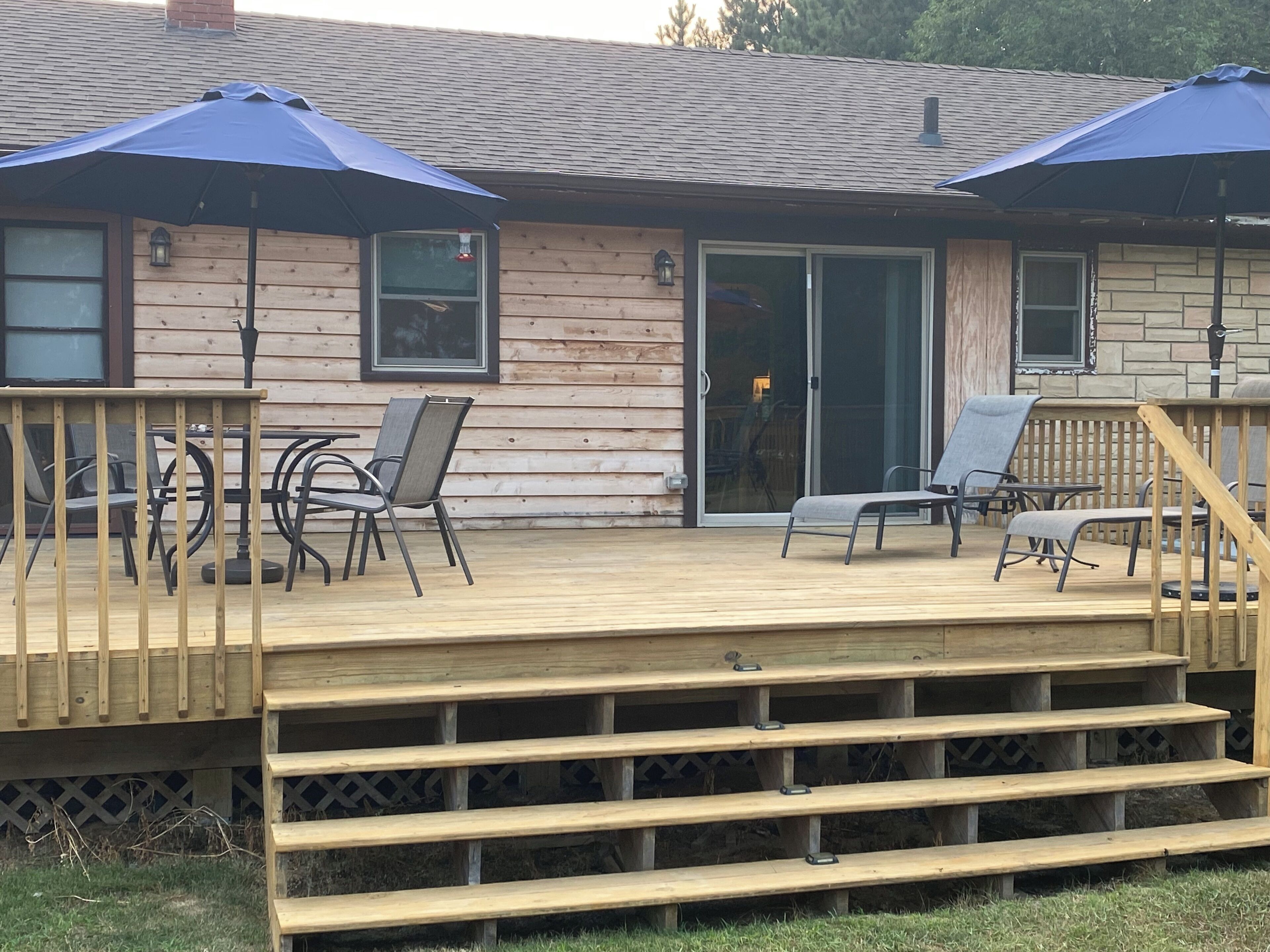 Una terraza de madera con mobiliario exterior y una escalera que lleva a un espacio cubierto con sombrillas.