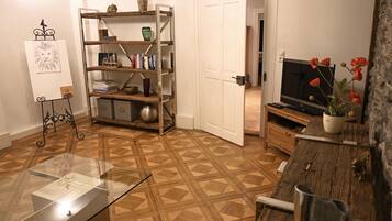 Ein Wohnzimmer mit Fischgrätparkett, einem Glastisch, einer Holzkommode, einem Fernseher und einem Bücherregal mit diversen Gegenständen.