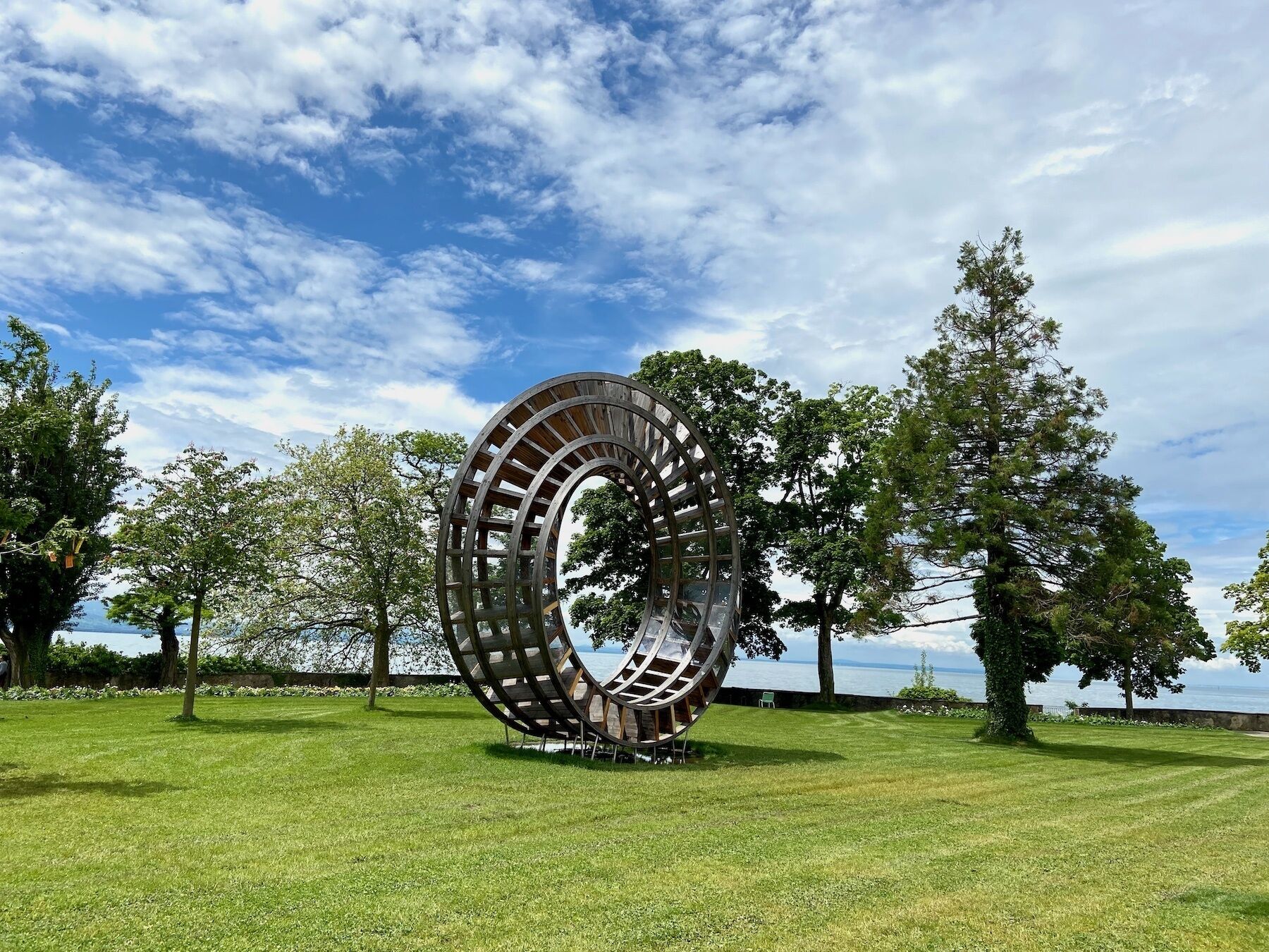 A large, circular metal sculpture in a grassy park area with trees and a body of water in the background.