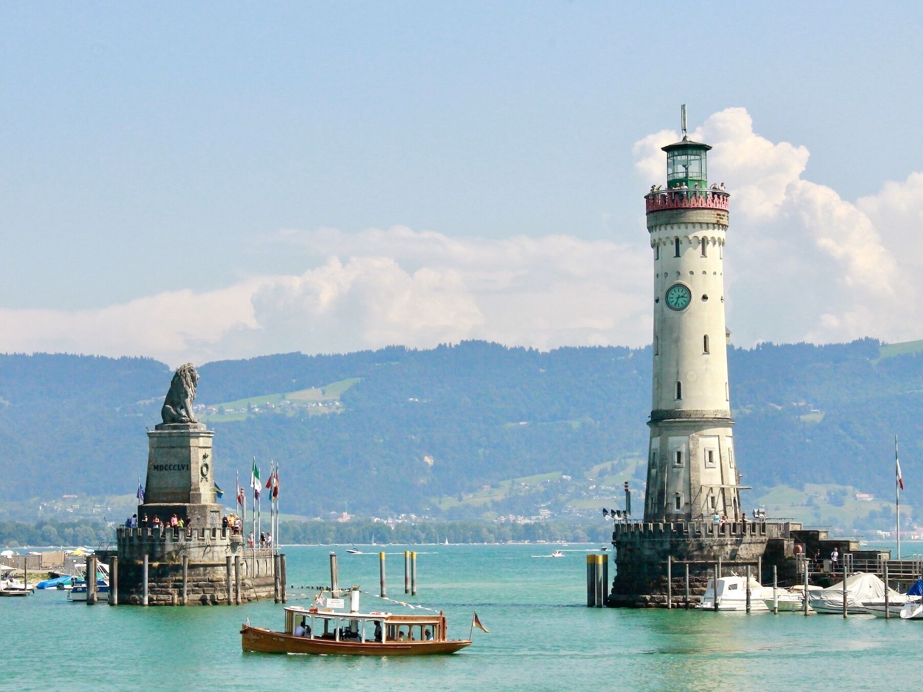 A lighthouse and a statue in a harbor with boats and a mountainous backdrop.