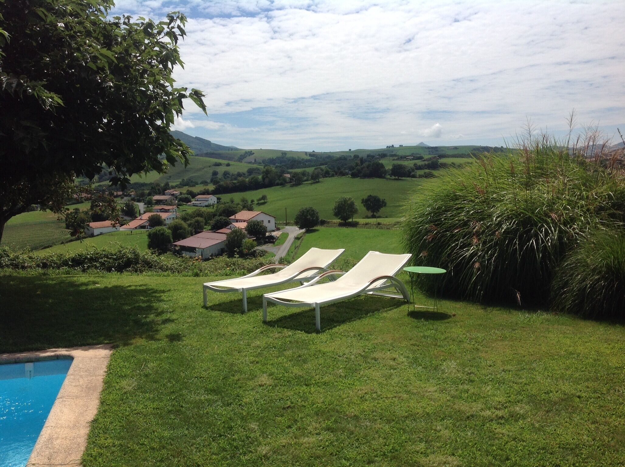 A poolside area with two lounge chairs and a small table, surrounded by lush greenery and a view of a rural landscape with houses and hills in the distance.