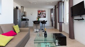 A modern living room with a glass coffee table, a sofa with colorful cushions, a kitchen area with a dining set, and a TV mounted on the wall. - CANNES CENTER, IDEAL CONGRESS, HOLIDAYS, MODERN APARTMENT FACING THE OLD PORT (Cannes)