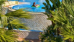 Una piscina con una persona en un flotador, rodeada de exuberante follaje y un edificio con tejado de baldosas.