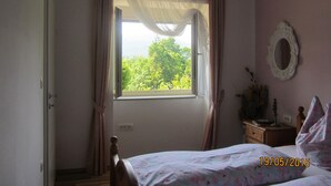 Un dormitorio con cama, una ventana con cortinas translúcidas, una mesita de noche con lámpara y un espejo en la pared.