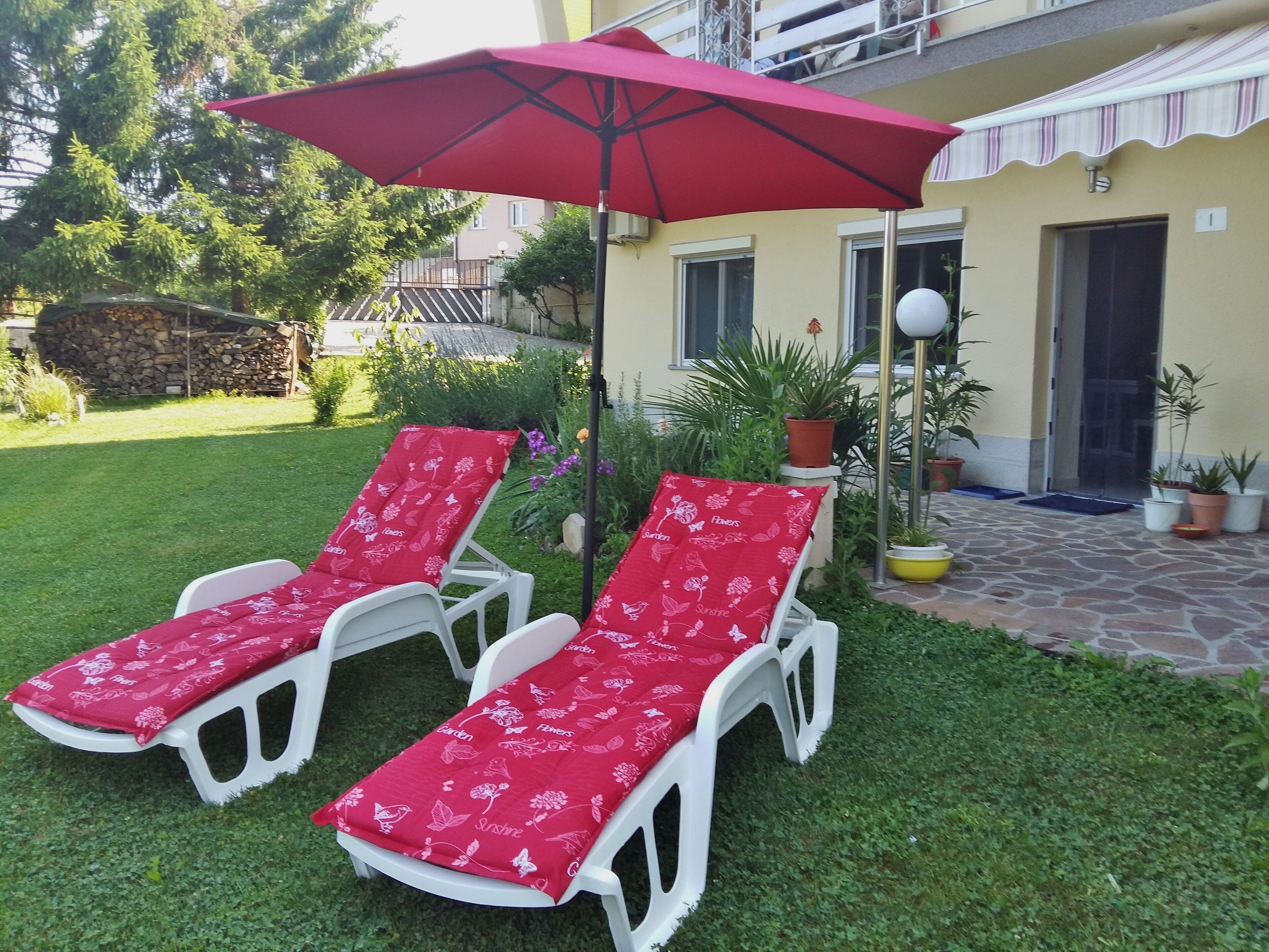 Two white lounge chairs with red floral cushions, a red umbrella, and potted plants in a garden area.