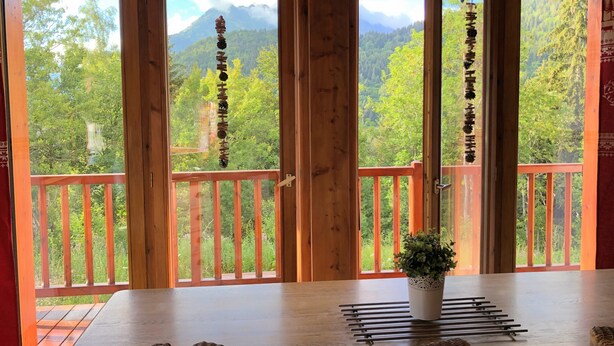 Ein Zimmer mit Holztisch und -stühlen, einer Topfpflanze und Blick auf einen bewaldeten Gebirgszug durch große Fenster.