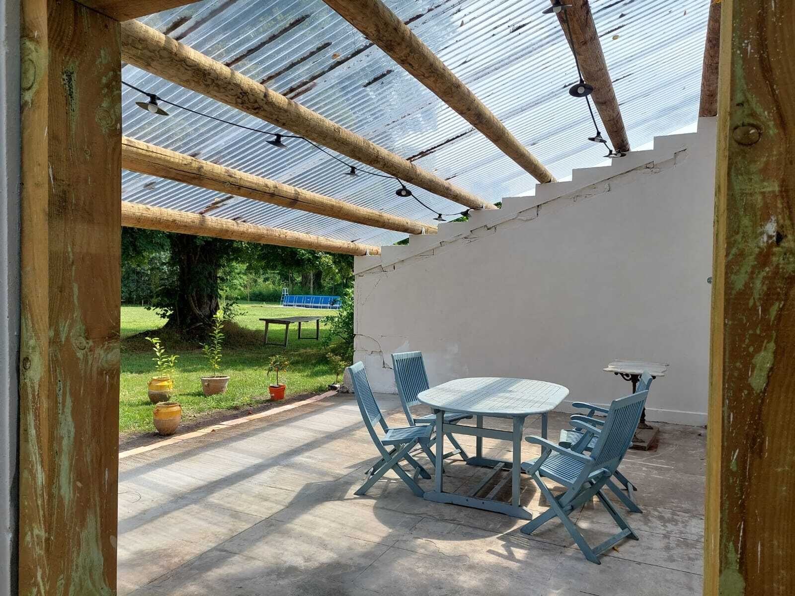 Eine überdachte Terrasse mit Tisch und Stühlen, ein überdachter Pergola und Blick auf eine grüne Landschaft.