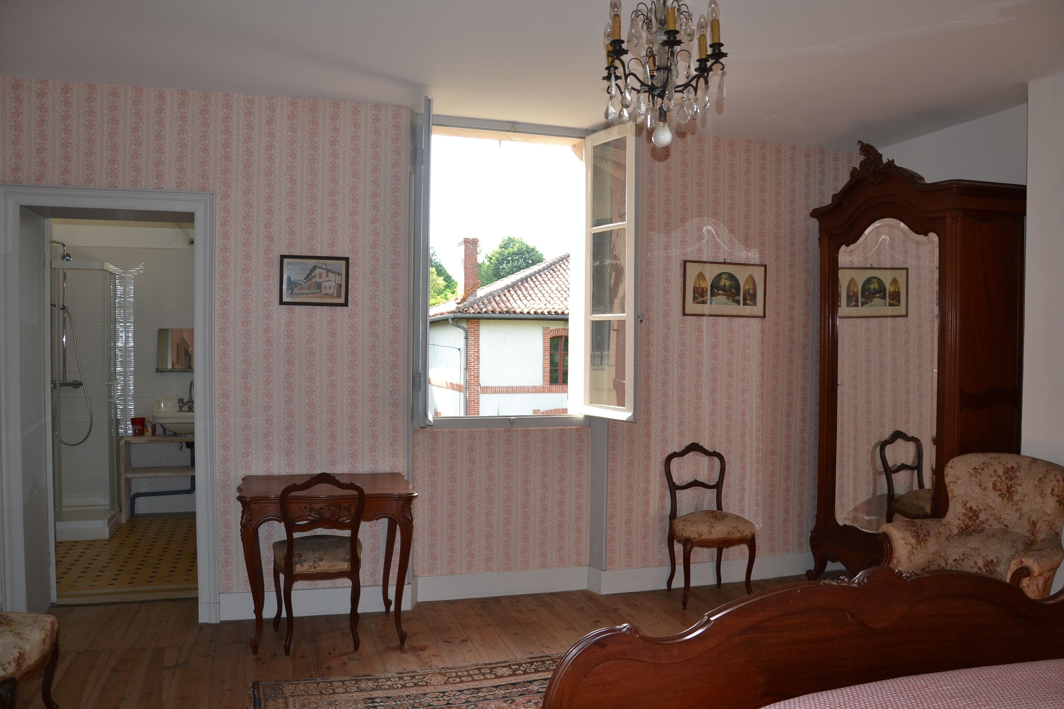 A bedroom with a wooden bed, two chairs, a small table, and a window with a view of a house.
